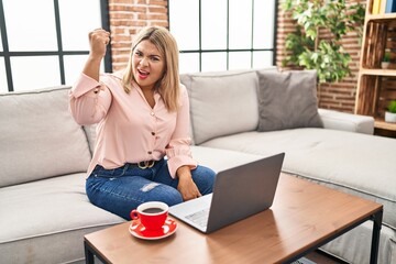 Young hispanic woman using laptop sitting on the sofa at home angry and mad raising fist frustrated and furious while shouting with anger. rage and aggressive concept.