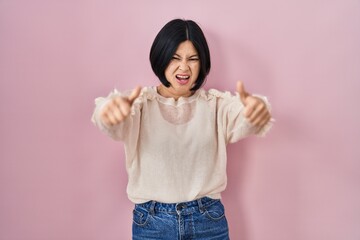 Young asian woman standing over pink background approving doing positive gesture with hand, thumbs up smiling and happy for success. winner gesture.