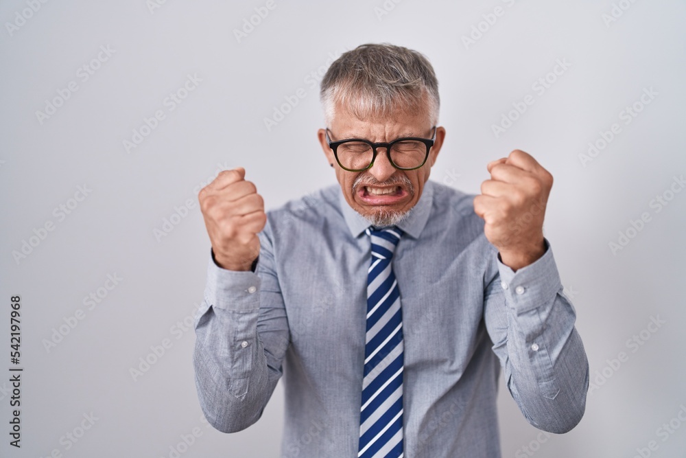 Sticker Hispanic business man with grey hair wearing glasses angry and mad raising fists frustrated and furious while shouting with anger. rage and aggressive concept.