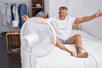 Middle age grey-haired man using ventilator sitting on bed at bedroom