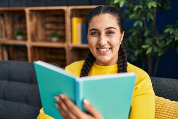 Fototapeta premium Young hispanic woman reading book sitting on sofa at home