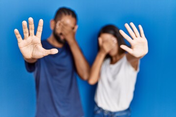 Young hispanic couple standing together covering eyes with hands and doing stop gesture with sad and fear expression. embarrassed and negative concept.