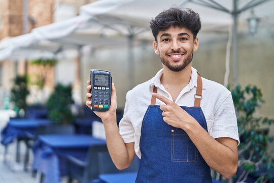 Arab man with beard wearing waiter apron at restaurant terrace holding dataphone smiling happy pointing with hand and finger