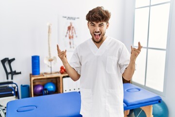 Young arab man working at pain recovery clinic shouting with crazy expression doing rock symbol with hands up. music star. heavy concept.