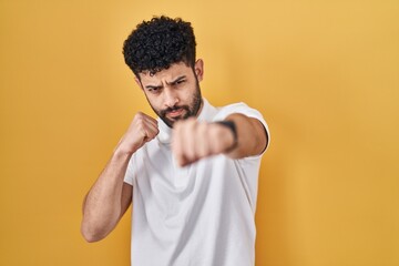 Arab man standing over yellow background punching fist to fight, aggressive and angry attack,...