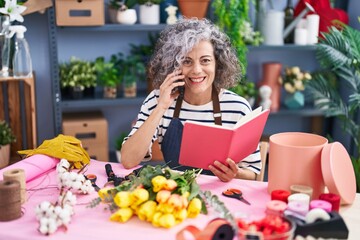 Middle age grey-haired woman florist talking on smartphone reading book at florist