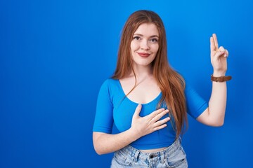 Fototapeta premium Redhead woman standing over blue background smiling swearing with hand on chest and fingers up, making a loyalty promise oath