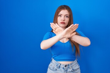 Redhead woman standing over blue background rejection expression crossing arms and palms doing negative sign, angry face