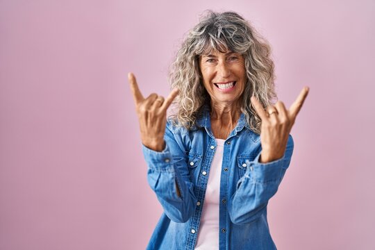 Middle age woman standing over pink background shouting with crazy expression doing rock symbol with hands up. music star. heavy concept.