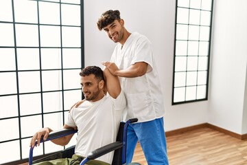 Two hispanic men physiotherapist and patient sitting on wheelchair having rehab session stretching arm at clinic