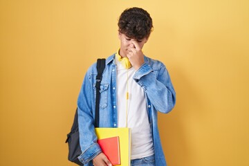 Hispanic teenager wearing student backpack and holding books tired rubbing nose and eyes feeling...