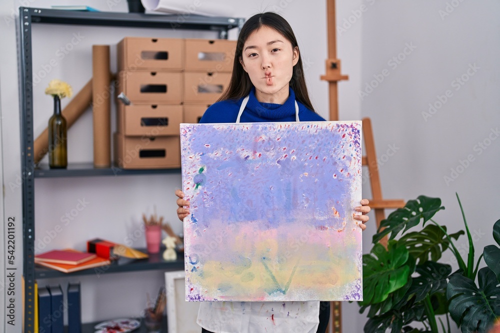 Sticker Chinese young woman holding canvas at art studio making fish face with mouth and squinting eyes, crazy and comical.