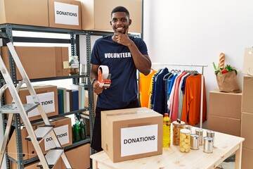 Young african american volunteer man packing donations box for charity with hand on chin thinking about question, pensive expression. smiling and thoughtful face. doubt concept.