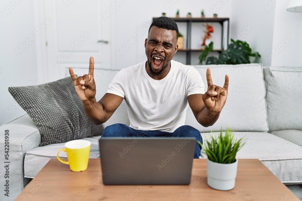 Sticker Young african man using laptop at home shouting with crazy expression doing rock symbol with hands up. music star. heavy concept.