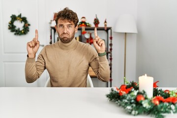 Young handsome man with beard sitting on the table by christmas decoration pointing up looking sad...