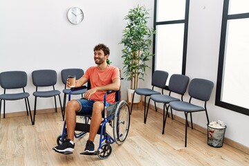 Young hispanic man drinking coffee sitting on wheelchair at clinic waiting room.