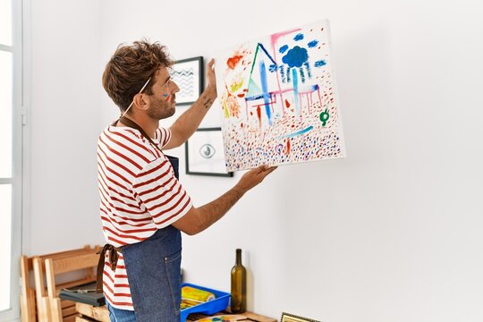 Young hispanic man hanging canvas on wall at art studio