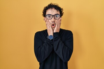 Fototapeta premium Hispanic man standing over yellow background afraid and shocked, surprise and amazed expression with hands on face