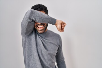 Hispanic man with beard standing over white background covering eyes with arm smiling cheerful and funny. blind concept.