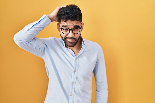 Hispanic man with beard standing over yellow background confuse and wonder about question. uncertain with doubt, thinking with hand on head. pensive concept.