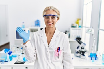 Middle age blonde woman working at scientist laboratory smiling cheerful presenting and pointing with palm of hand looking at the camera.