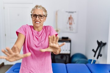 Middle age blonde woman at pain recovery clinic afraid and terrified with fear expression stop gesture with hands, shouting in shock. panic concept.