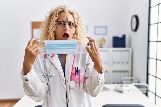 Middle age blonde woman wearing doctor uniform and medical mask in shock face, looking skeptical and sarcastic, surprised with open mouth