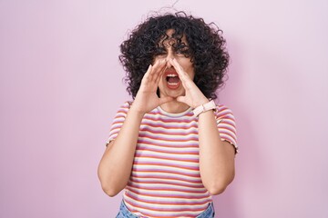 Young middle east woman standing over pink background shouting angry out loud with hands over mouth