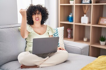 Young brunette woman with curly hair using laptop sitting on the sofa at home angry and mad raising fist frustrated and furious while shouting with anger. rage and aggressive concept.