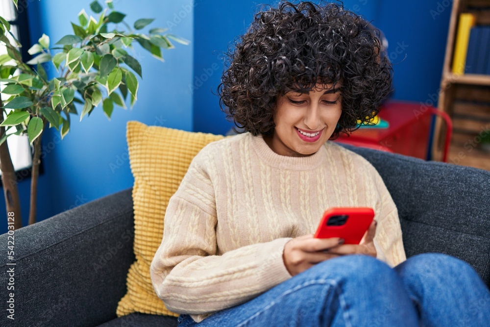 Sticker young middle east woman using smartphone sitting on sofa at home