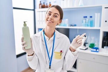 Young caucasian woman working at scientist laboratory holding body lotion smiling happy pointing with hand and finger to the side
