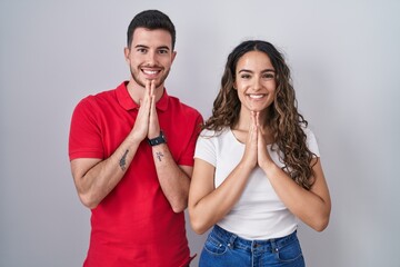 Young hispanic couple standing over isolated background praying with hands together asking for forgiveness smiling confident.