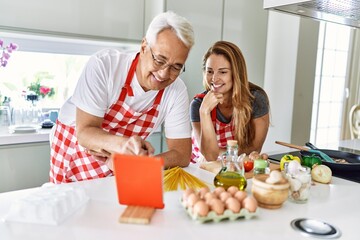 Middle age hispanic couple smiling happy cooking and using touchpad at the kitchen.