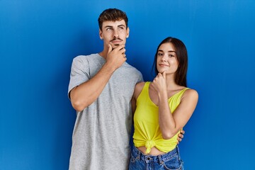 Fototapeta premium Young hispanic couple standing together over blue background looking confident at the camera smiling with crossed arms and hand raised on chin. thinking positive.
