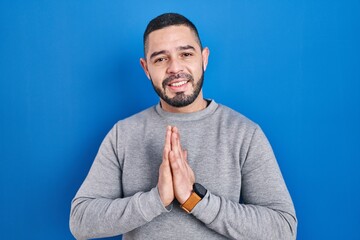 Hispanic man standing over blue background praying with hands together asking for forgiveness smiling confident.