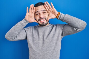 Hispanic man standing over blue background smiling cheerful playing peek a boo with hands showing face. surprised and exited