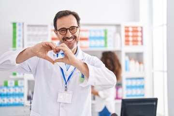 Middle age hispanic man working at pharmacy drugstore smiling in love doing heart symbol shape with...