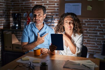 Middle age hispanic couple using touchpad sitting on the table at night pointing aside worried and...