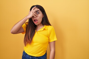 Young arab woman standing over yellow background peeking in shock covering face and eyes with hand,...