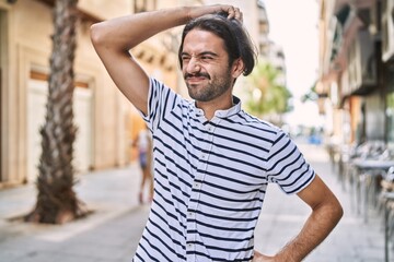 Young hispanic man with beard outdoors at the city confuse and wonder about question. uncertain with doubt, thinking with hand on head. pensive concept.