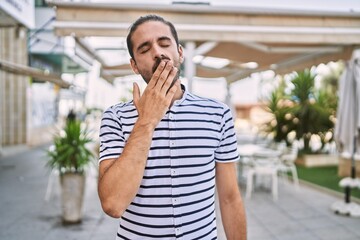 Young hispanic man with beard outdoors at the city bored yawning tired covering mouth with hand....