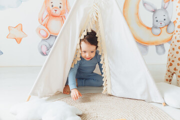 Fototapeta na wymiar A children's room with a tent-wigwam. the kid is playing in a white sunny bedroom. Cozy bright interior for a children's room. A little boy with toy cubes is playing on a wooden floor.
