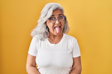 Middle age woman with grey hair standing over yellow background sticking tongue out happy with funny expression. emotion concept.