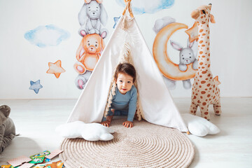 A children's room with a tent-wigwam. the kid is playing in a white sunny bedroom. Cozy bright interior for a children's room. A little boy with toy cubes is playing on a wooden floor. © inna717