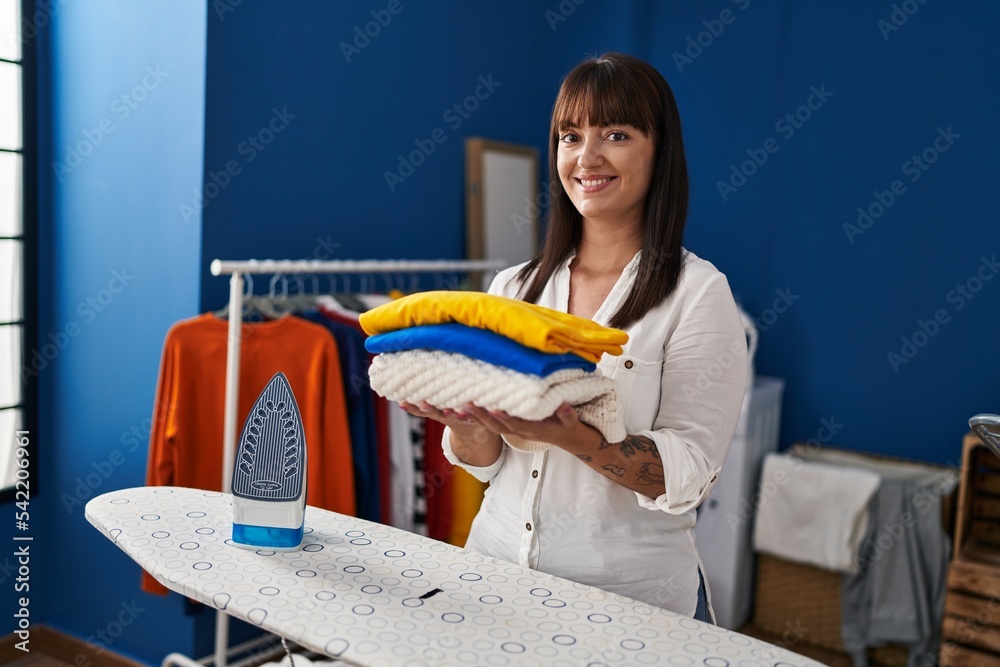 Wall mural young beautiful hispanic woman smiling confident holding folded clothes at laundry room - Wall murals