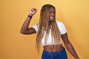 African american woman with braided hair standing over yellow background dancing happy and...