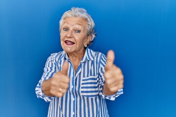 Senior woman with grey hair standing over blue background approving doing positive gesture with hand, thumbs up smiling and happy for success. winner gesture.