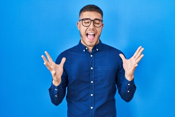 Young hispanic man wearing glasses over blue background celebrating mad and crazy for success with...