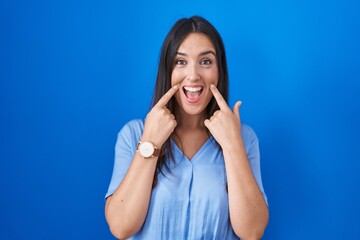 Young brunette woman standing over blue background smiling with open mouth, fingers pointing and forcing cheerful smile