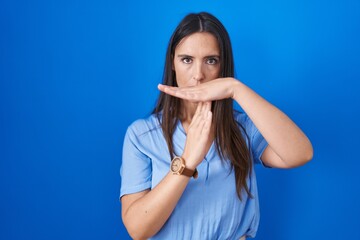 Young brunette woman standing over blue background doing time out gesture with hands, frustrated and serious face